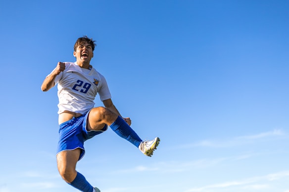 A soccer player is caught in mid-air against a clear blue sky, wearing a white jersey with the number 29 and blue shorts. The expression on the player