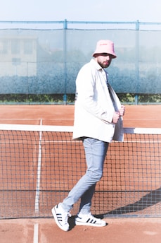 A man stands confidently on a tennis court, leaning slightly against the net. He is wearing a white jacket, pink bucket hat, and gray pants, paired with white and black Adidas sneakers. The background features a blue mesh fence and a blurred building.