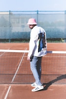 A man stands on a clay tennis court, next to the net. He is wearing a pink bucket hat, a white jacket with a colorful pattern on the back, gray pants, and white shoes with black stripes. The background includes a fence and a blurry view of buildings.