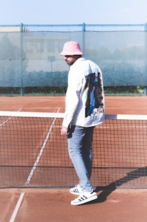 A man stands on a clay tennis court, next to the net. He is wearing a pink bucket hat, a white jacket with a colorful pattern on the back, gray pants, and white shoes with black stripes. The background includes a fence and a blurry view of buildings.