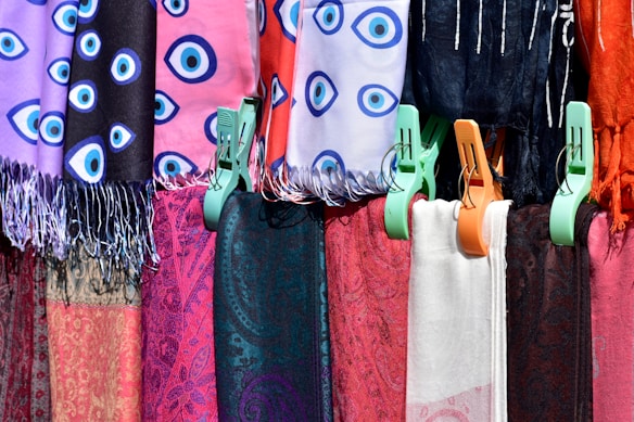 A collection of colorful fabrics hang side by side, each attached with plastic clips. Some of the fabrics have eye-like patterns in blue, others have intricate paisley designs. The colors range from purples and reds to blacks and whites.