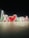 The image depicts illuminated giant letters forming the phrase 'I ❤️ UAE' against a dark background. The letters are lit up with white outlines, and the colors red and green are prominently featured. There are three people standing in front of the display, casting long shadows on the tiled ground.