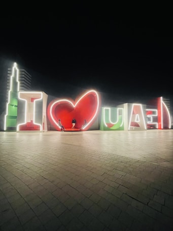 The image depicts illuminated giant letters forming the phrase 'I ❤️ UAE' against a dark background. The letters are lit up with white outlines, and the colors red and green are prominently featured. There are three people standing in front of the display, casting long shadows on the tiled ground.