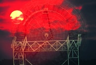 A large radar dish structure is silhouetted against a dramatic red and black sky. The setting or rising sun is visibly bright, partially obscured by clouds, creating a striking contrast with the dark metallic structure.