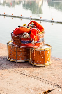 A set of weathered drums sits stacked on a stone surface near a body of water. A colorful fabric and pipes, possibly part of a bagpipe, rest atop the drums. The surface shows signs of age and wear, while the water in the background reflects serene light.