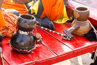 A vibrant scene features musical instruments including a gourd drum and a bell, placed on a red wooden surface. The background shows colorful fabrics in shades of orange, yellow, and patterned red, along with a small wooden mallet. The arrangement suggests a cultural or traditional setup.