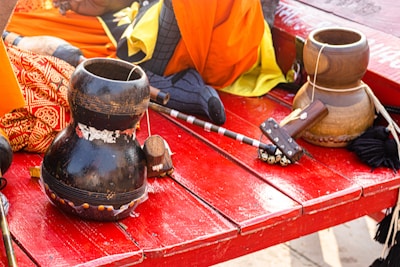 A vibrant scene features musical instruments including a gourd drum and a bell, placed on a red wooden surface. The background shows colorful fabrics in shades of orange, yellow, and patterned red, along with a small wooden mallet. The arrangement suggests a cultural or traditional setup.