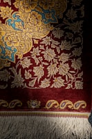 A richly woven carpet featuring intricate floral patterns in shades of yellow and blue on a deep red background. The lower edge displays decorative tassels.