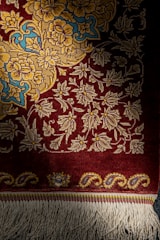 A richly woven carpet featuring intricate floral patterns in shades of yellow and blue on a deep red background. The lower edge displays decorative tassels.