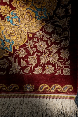 A richly woven carpet featuring intricate floral patterns in shades of yellow and blue on a deep red background. The lower edge displays decorative tassels.