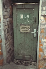 A weathered green door set in a brick wall, featuring an emergency exit sign with a running figure and arrow. The door has noticeable wear and rust, with a metal handle and a sign in German warning that entry is forbidden to unauthorized persons.