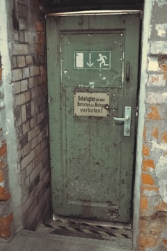 A weathered green door set in a brick wall, featuring an emergency exit sign with a running figure and arrow. The door has noticeable wear and rust, with a metal handle and a sign in German warning that entry is forbidden to unauthorized persons.