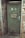 A weathered green door set in a brick wall, featuring an emergency exit sign with a running figure and arrow. The door has noticeable wear and rust, with a metal handle and a sign in German warning that entry is forbidden to unauthorized persons.