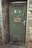 A weathered green door set in a brick wall, featuring an emergency exit sign with a running figure and arrow. The door has noticeable wear and rust, with a metal handle and a sign in German warning that entry is forbidden to unauthorized persons.
