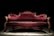 A luxurious Victorian-style sofa with elegant, deep red velvet upholstery and ornate wooden carvings. The setting is dramatically lit, highlighting the intricate textures and curves of the sofa against a dark background.