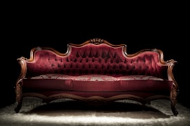 A luxurious Victorian-style sofa with elegant, deep red velvet upholstery and ornate wooden carvings. The setting is dramatically lit, highlighting the intricate textures and curves of the sofa against a dark background.