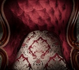 An ornate and luxurious piece of furniture with deep red upholstery and intricate gold patterns. The fabric features a tufted design, accented by wooden armrests with elaborate carvings.