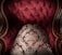 An ornate and luxurious piece of furniture with deep red upholstery and intricate gold patterns. The fabric features a tufted design, accented by wooden armrests with elaborate carvings.