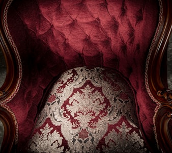 An ornate and luxurious piece of furniture with deep red upholstery and intricate gold patterns. The fabric features a tufted design, accented by wooden armrests with elaborate carvings.
