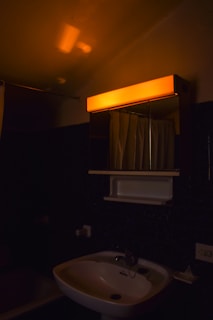 A dimly lit bathroom featuring a white sink with a dark faucet. Above the sink is a mirrored medicine cabinet with an orange light glowing at the top. A white shower curtain is visible behind the sink.