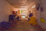 A narrow indoor garage or storage space with various items scattered throughout. On the left, there are stacked crates and a wheelbarrow, while a table draped in fabric holds more objects. On the right, clothes hang on a rack, and a large object is covered with a yellow cloth. A bicycle wheel and various bags are hung on the wall. Sunlight filters from the entrance, illuminating a garden gate and chairs outside.