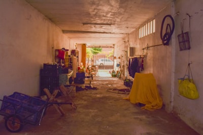 A narrow indoor garage or storage space with various items scattered throughout. On the left, there are stacked crates and a wheelbarrow, while a table draped in fabric holds more objects. On the right, clothes hang on a rack, and a large object is covered with a yellow cloth. A bicycle wheel and various bags are hung on the wall. Sunlight filters from the entrance, illuminating a garden gate and chairs outside.