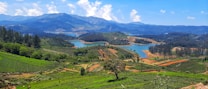 A breathtaking landscape featuring rolling green hills, a serene blue lake, and distant mountain ranges beneath a vibrant blue sky with scattered clouds. The area is lush with vegetation, including patches of dense forest and cultivated fields.