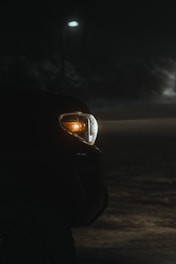 A vehicle's front light is illuminated during nighttime in a dimly lit environment. The scene is partially illuminated by a solitary streetlamp in the background, casting soft light over the dark surroundings.