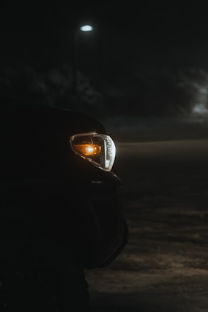 A vehicle's front light is illuminated during nighttime in a dimly lit environment. The scene is partially illuminated by a solitary streetlamp in the background, casting soft light over the dark surroundings.