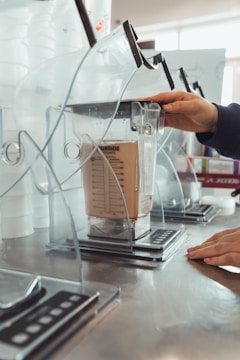 A close-up of a hand operating a commercial blender, with the appliance enclosed in a transparent protective cover. The blender jar is filled with a beige liquid, sitting on a metallic counter in an industrial-style setting.