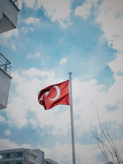 A Turkish flag flutters on a pole against a backdrop of a bright, partly cloudy sky. In the foreground, there are parts of a building with balconies and bare tree branches.