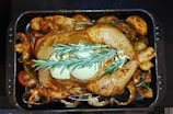 A whole roasted chicken placed in a black baking tray, garnished with sprigs of rosemary and pats of butter on top. Surrounding the chicken are various cooked vegetables, including mushrooms and sliced tomatoes, all seasoned and roasted.