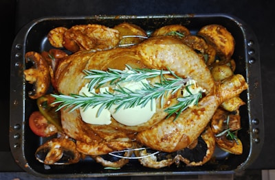 A whole roasted chicken placed in a black baking tray, garnished with sprigs of rosemary and pats of butter on top. Surrounding the chicken are various cooked vegetables, including mushrooms and sliced tomatoes, all seasoned and roasted.
