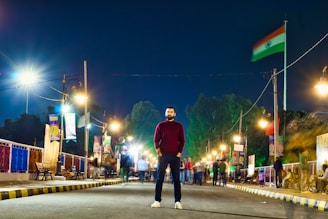A man stands confidently in the middle of a brightly lit street at night. There are streetlights on both sides of the road, banners, and an Indian flag on a pole. The background shows trees and blurred motion of people walking, suggesting a festive or busy atmosphere.