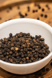 A white ceramic bowl filled with whole black peppercorns sits on a wooden surface. Some peppercorns have spilled out of the bowl and are scattered on the surface in the background.