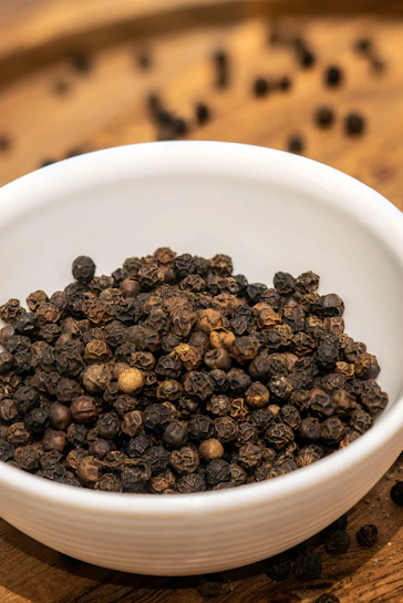 Fresh black peppercorns spilling from a small terracotta jar onto a white surface.