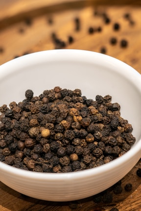 A white ceramic bowl filled with whole black peppercorns sits on a wooden surface. Some peppercorns have spilled out of the bowl and are scattered on the surface in the background.