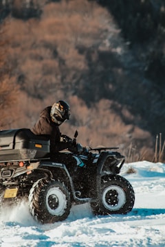 A person wearing a helmet rides an all-terrain vehicle (ATV) off-road in a snowy environment. The background features blurred trees, creating a dynamic and adventurous scene.