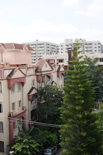 A residential area with a mix of modern and traditional architectural styles. High-rise apartment buildings in the background contrast with the older, beige and pink houses with sloped roofs in the foreground. Lush greenery, including a tall coniferous tree, surrounds the buildings, adding to the suburban feel.