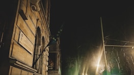 A nighttime scene featuring a historic building with an arched entrance and a hanging flag with stars. An illuminated plaque is visible on the wall. The photo is dimly lit with streetlights casting a warm glow.