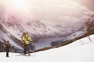Two people are trekking through a snow-covered mountain landscape, carrying ski poles and backpacks. The scene is set against a backdrop of rugged, snow-capped peaks and a distant view of a fjord or river. The sun shines through a cloudy sky, casting a soft, warm light across the scene.