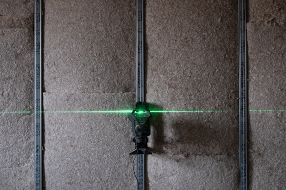 A green laser level projects a horizontal line across a textured wall, with vertically aligned metal studs visible. The scene suggests a construction or renovation setting.