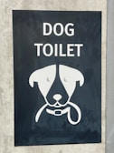 A sign with an illustration of a dog's face, featuring simplistic and stylized features including a collar. Above the illustration, the words 'DOG TOILET' are printed in bold, white letters. The sign is set on a dark background mounted on a concrete surface.
