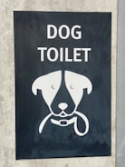 A sign with an illustration of a dog's face, featuring simplistic and stylized features including a collar. Above the illustration, the words 'DOG TOILET' are printed in bold, white letters. The sign is set on a dark background mounted on a concrete surface.