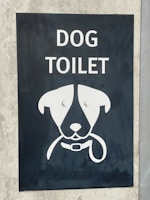 A sign with an illustration of a dog's face, featuring simplistic and stylized features including a collar. Above the illustration, the words 'DOG TOILET' are printed in bold, white letters. The sign is set on a dark background mounted on a concrete surface.