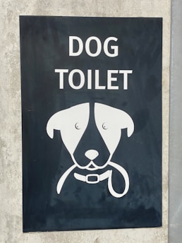 A sign with an illustration of a dog's face, featuring simplistic and stylized features including a collar. Above the illustration, the words 'DOG TOILET' are printed in bold, white letters. The sign is set on a dark background mounted on a concrete surface.