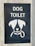 A sign with an illustration of a dog's face, featuring simplistic and stylized features including a collar. Above the illustration, the words 'DOG TOILET' are printed in bold, white letters. The sign is set on a dark background mounted on a concrete surface.