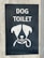 A sign with an illustration of a dog's face, featuring simplistic and stylized features including a collar. Above the illustration, the words 'DOG TOILET' are printed in bold, white letters. The sign is set on a dark background mounted on a concrete surface.