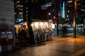 An outdoor food stall on a city street corner, illuminated by warm, hanging lights and sheltering a few patrons seated inside. A tree partially obscures the view, and there is a red post box on the sidewalk. The surrounding area is dimly lit, with tall buildings and street signage visible in the background.