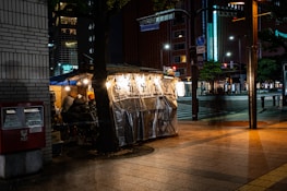 An outdoor food stall on a city street corner, illuminated by warm, hanging lights and sheltering a few patrons seated inside. A tree partially obscures the view, and there is a red post box on the sidewalk. The surrounding area is dimly lit, with tall buildings and street signage visible in the background.
